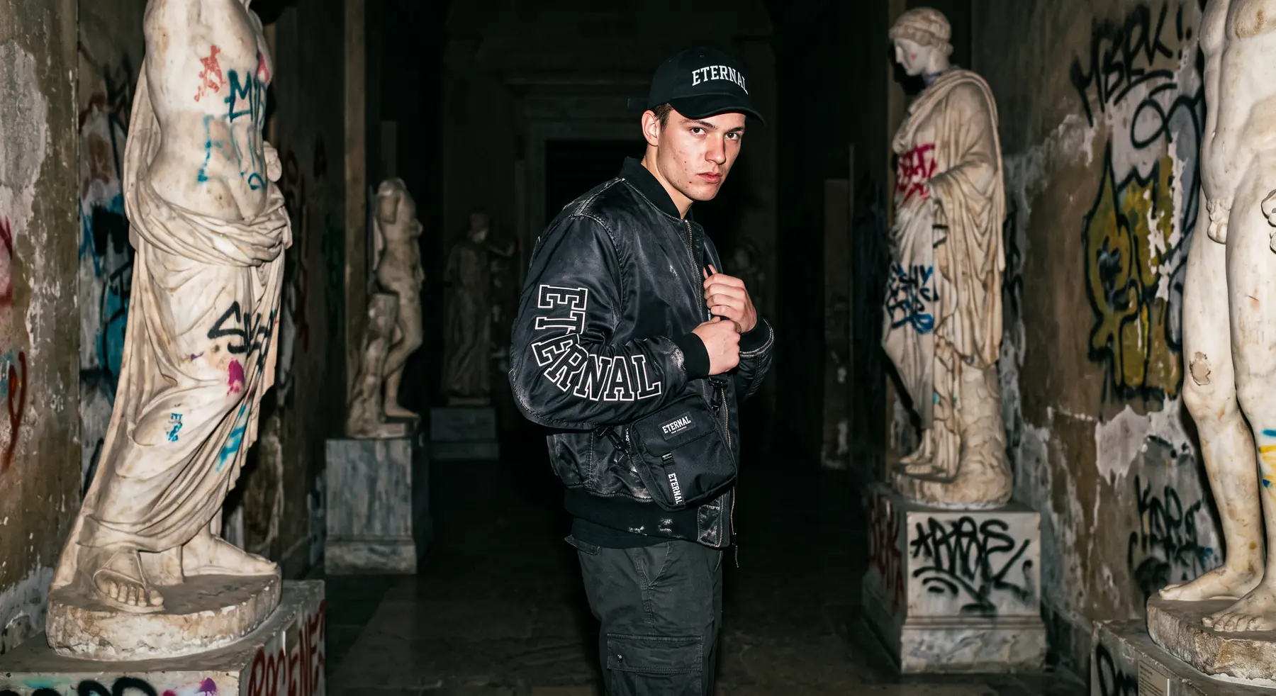 Hombre con chaqueta bomber de Eternal en un pasillo con estatuas y grafitis