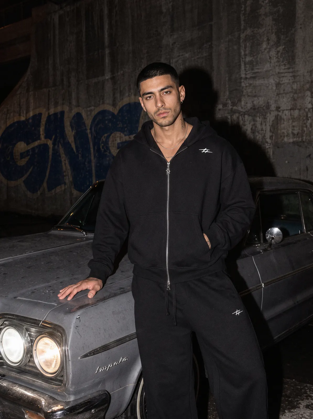 Modelo con chándal negro de GNG apoyado en un coche clásico de noche